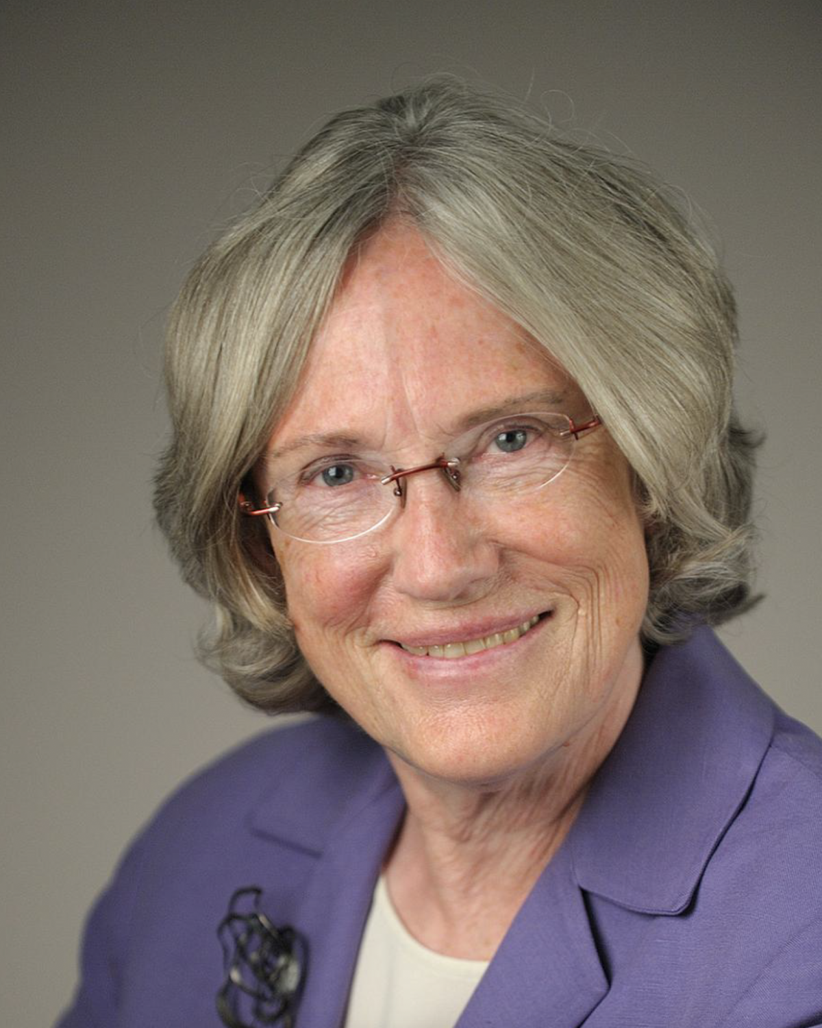 An older woman with short gray hair and glasses smiles at the camera. She is wearing a purple blazer over a light top with a brooch pinned to her lapel. The background is neutral and softly lit.