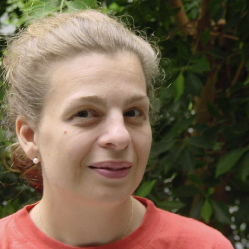 A woman with light skin and brown hair pulled back smiles slightly while wearing a red shirt. She is outdoors with green foliage in the background.