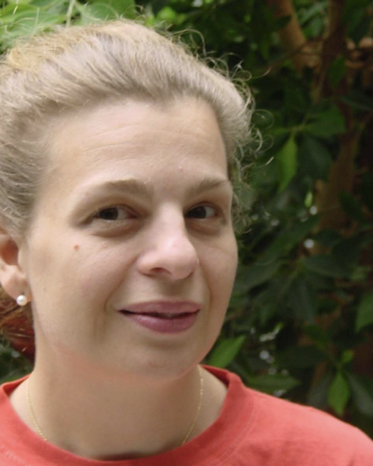 A woman with light skin and brown hair pulled back smiles slightly while wearing a red shirt. She is outdoors with green foliage in the background.