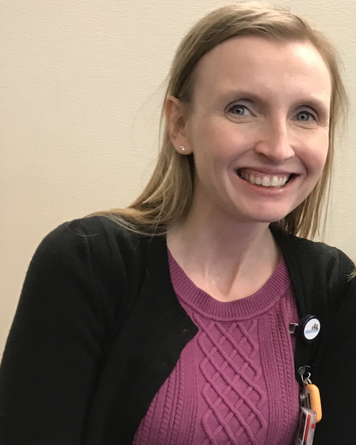 A woman with light hair, wearing a pink sweater and black cardigan, smiles at the camera. She has light skin and is sitting indoors against a beige wall.