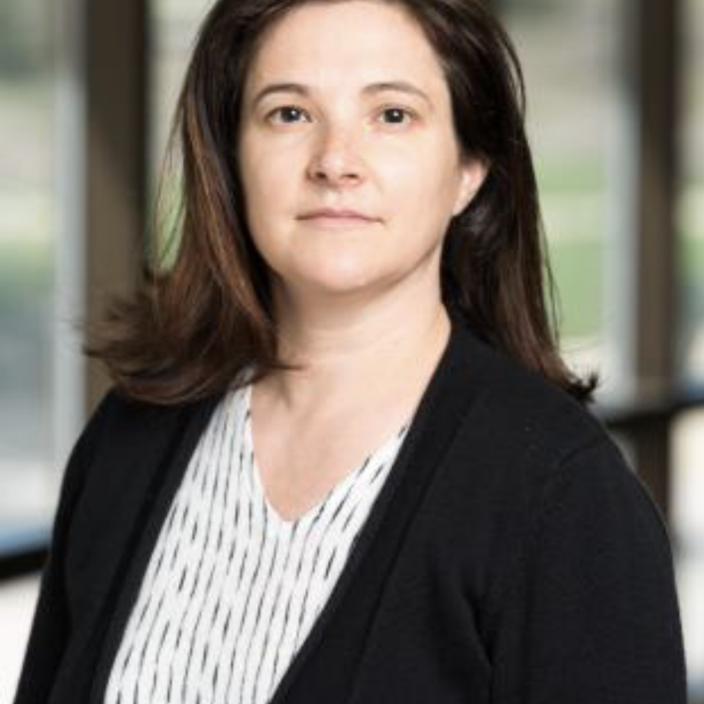 A woman with shoulder-length brown hair wearing a black cardigan and a white patterned blouse stands indoors with large windows and greenery in the background.