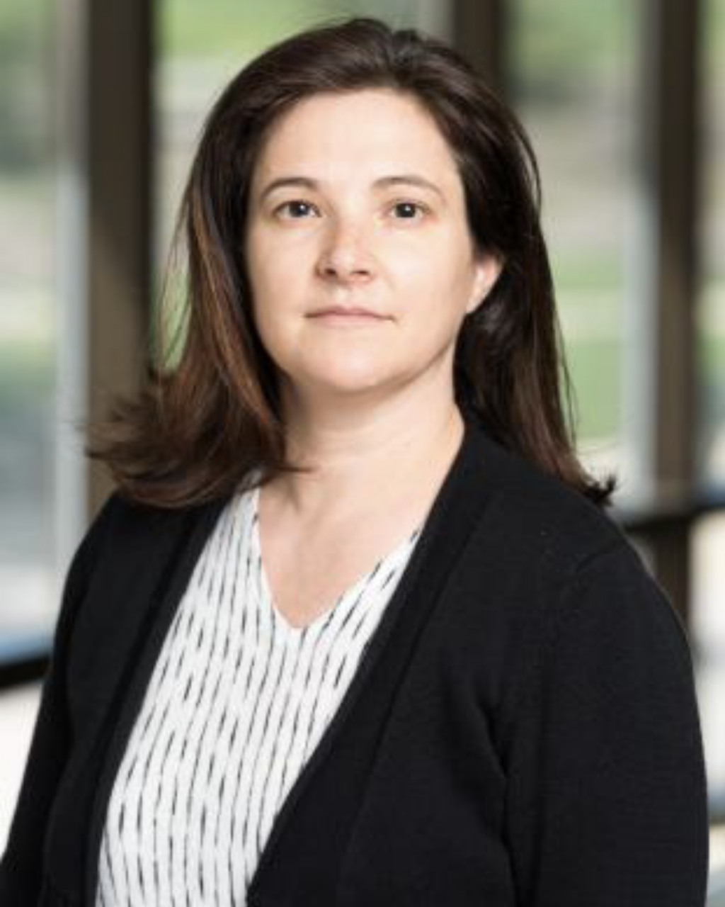 A woman with shoulder-length brown hair wearing a black cardigan and a white patterned blouse stands indoors with large windows and greenery in the background.