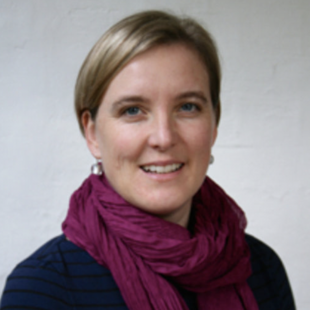 A woman with short blonde hair smiles at the camera. She is wearing a dark blue top and a purple scarf. The background is plain and light-colored.