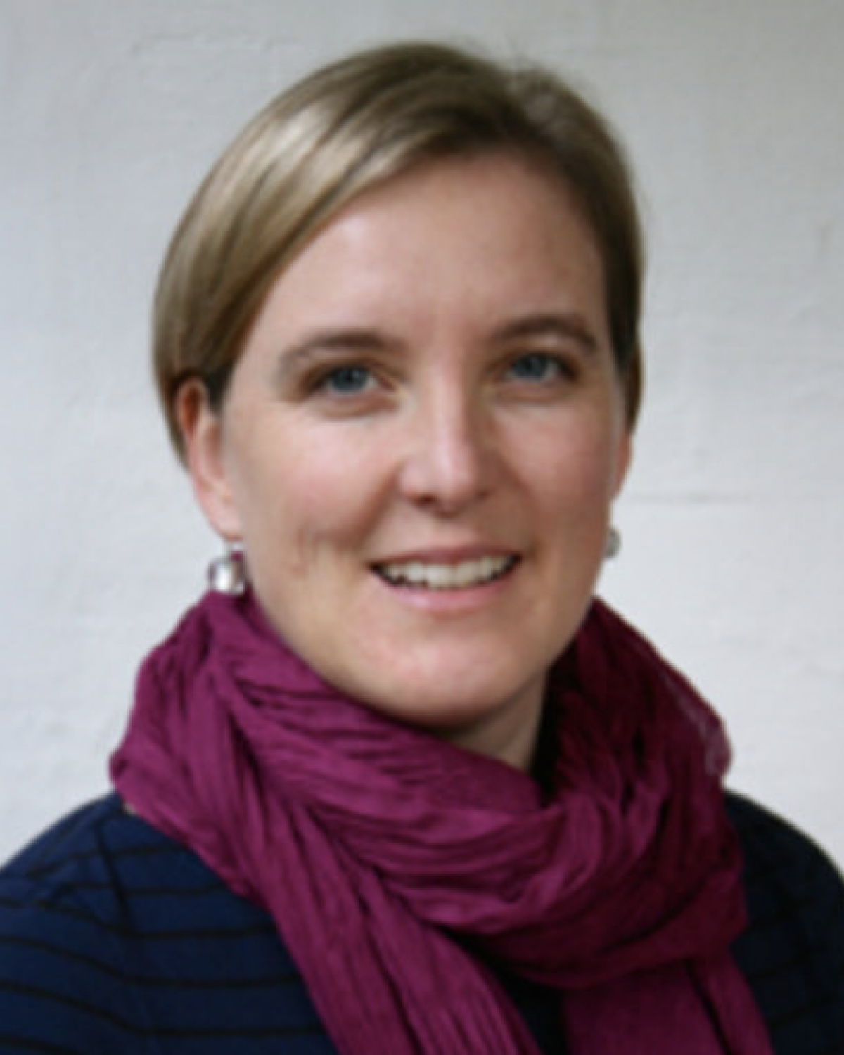 A woman with short blonde hair smiles at the camera. She is wearing a dark blue top and a purple scarf. The background is plain and light-colored.
