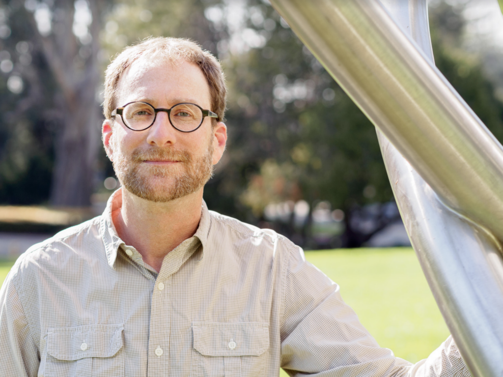 A man with short hair, glasses, and a beard stands outdoors by a large metal sculpture, wearing a light-colored button-up shirt. The background features green grass and trees.