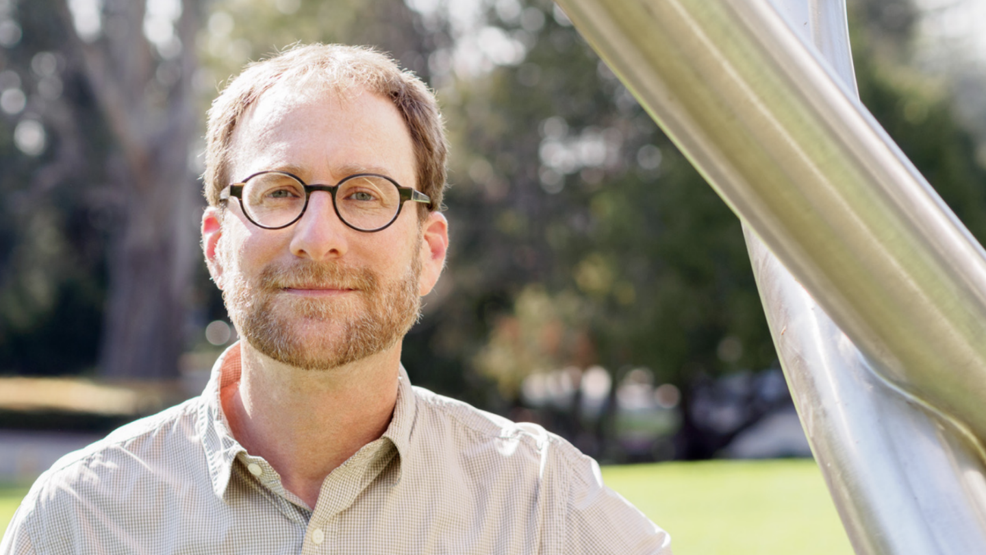 A man with short brown hair, a beard, and glasses stands outdoors near a large metal sculpture, with green grass and trees blurred in the background.