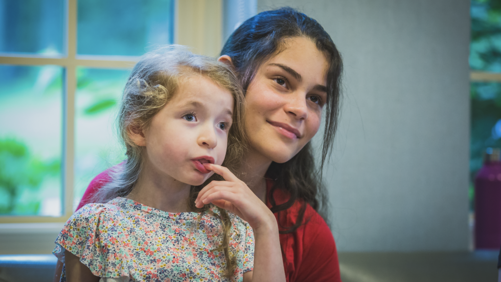 A young woman smiles while sitting with a little girl in a floral dress, who looks thoughtful with her finger on her lips. They are indoors with a window in the background.