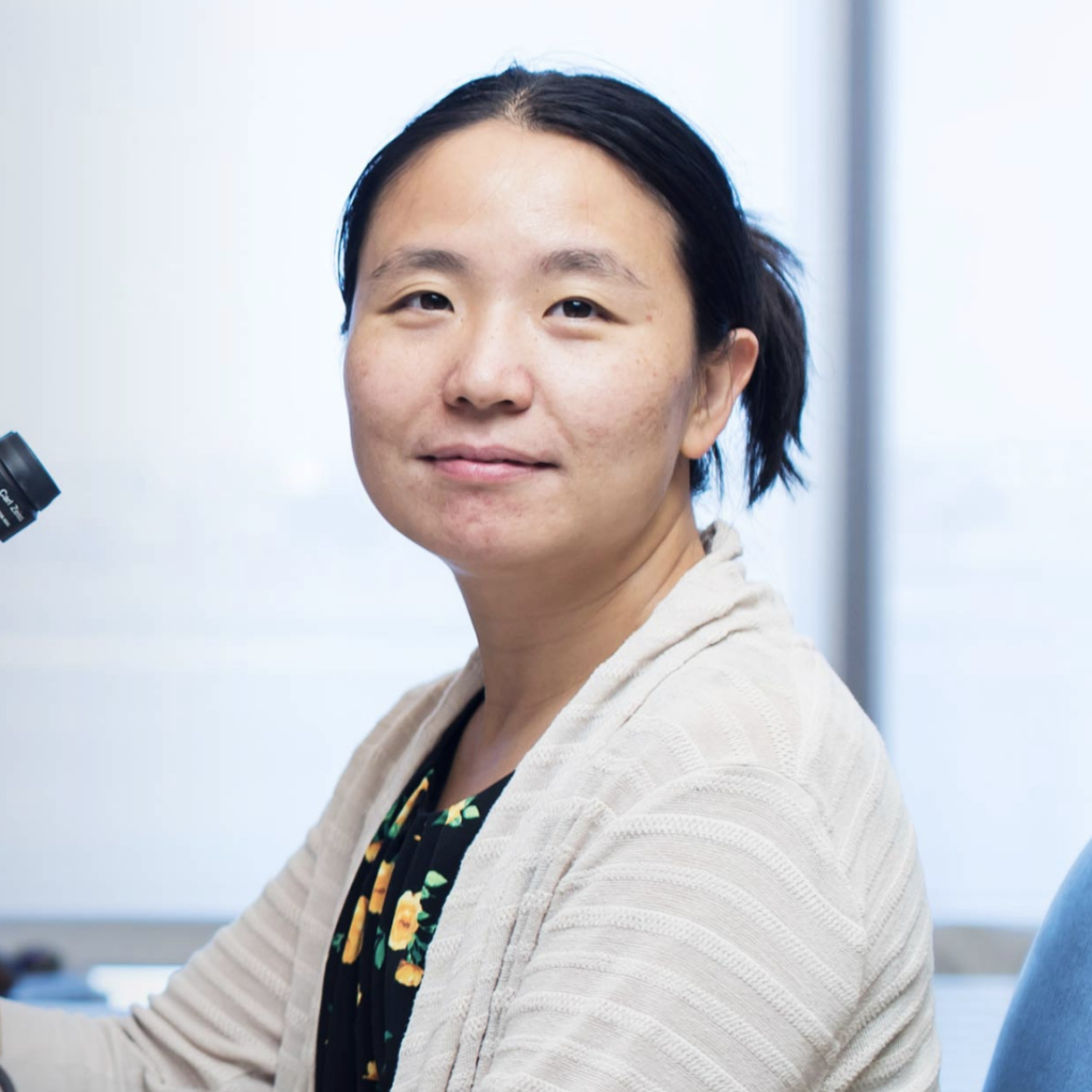 A woman with dark hair tied back, wearing a light striped cardigan over a floral top, sits at a desk next to a microscope and smiles gently at the camera in a bright, modern room.
