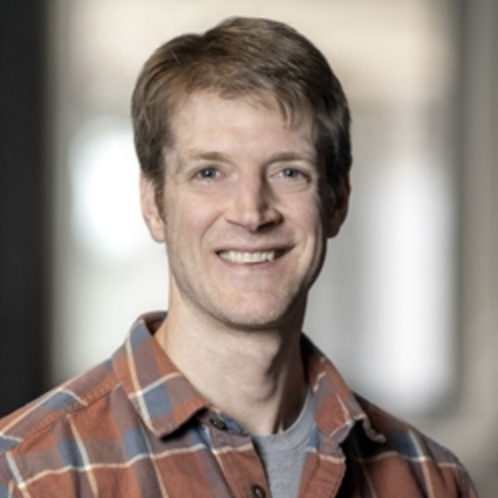 A smiling man with short brown hair wears a plaid shirt over a gray t-shirt, standing indoors with a blurred background.