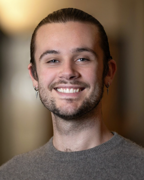 A young person with light skin, brown hair in a bun, and facial hair smiles at the camera, wearing a gray sweater and small hoop earrings, with a softly blurred indoor background.