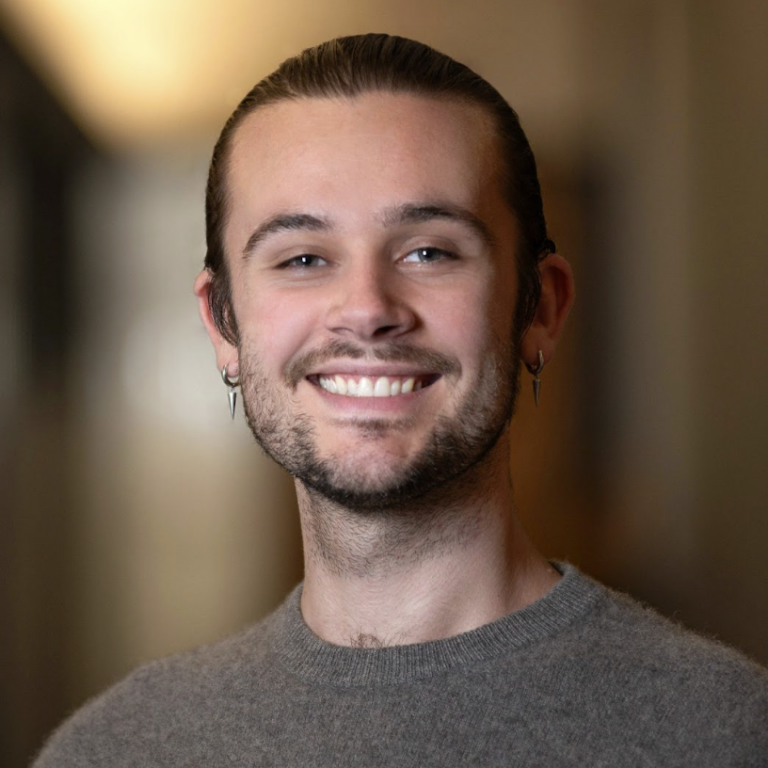 A young person with light skin, brown hair in a bun, and facial hair smiles at the camera, wearing a gray sweater and small hoop earrings, with a softly blurred indoor background.