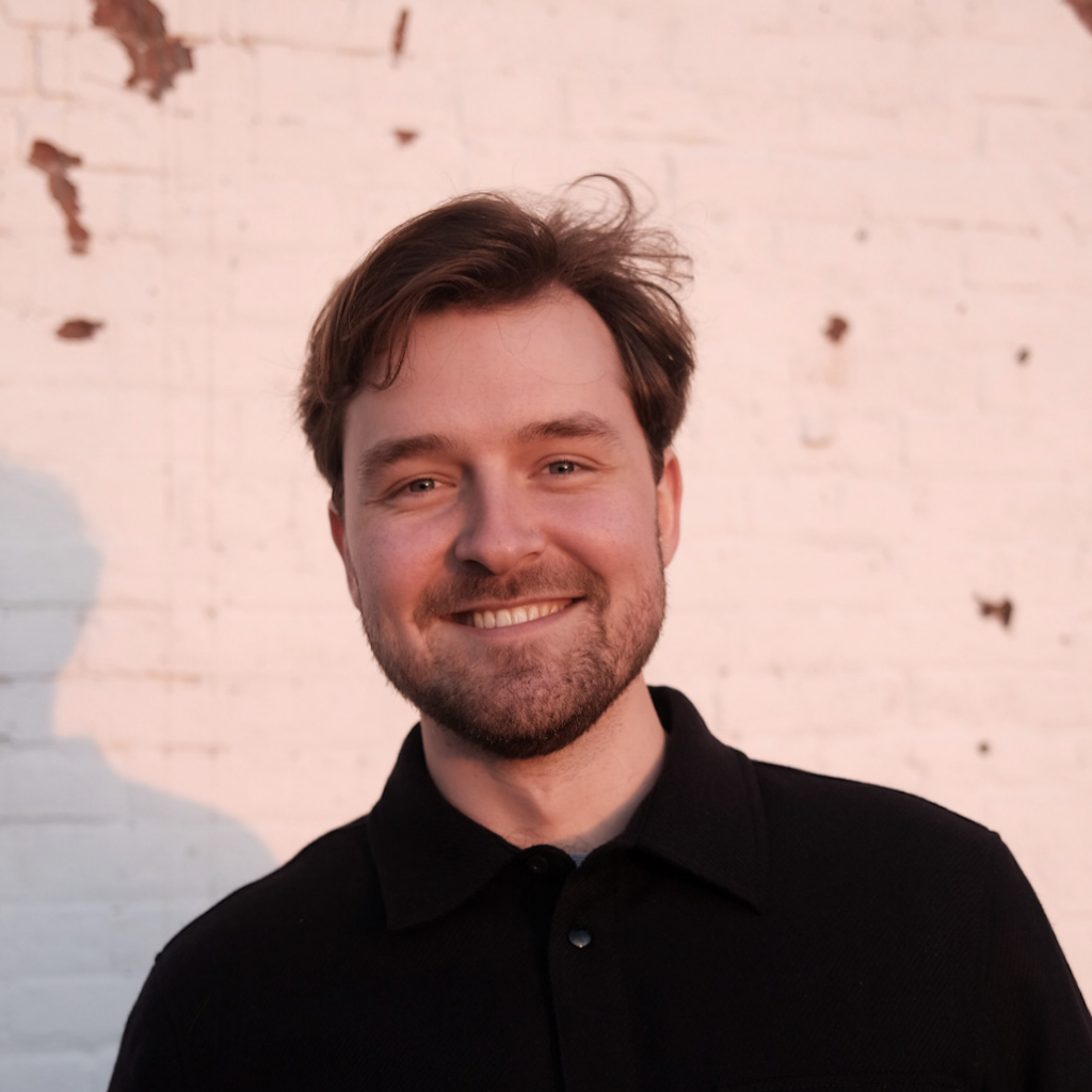 A man with brown hair and a beard smiles while standing in front of a white brick wall with some chipped paint, illuminated by warm sunlight.