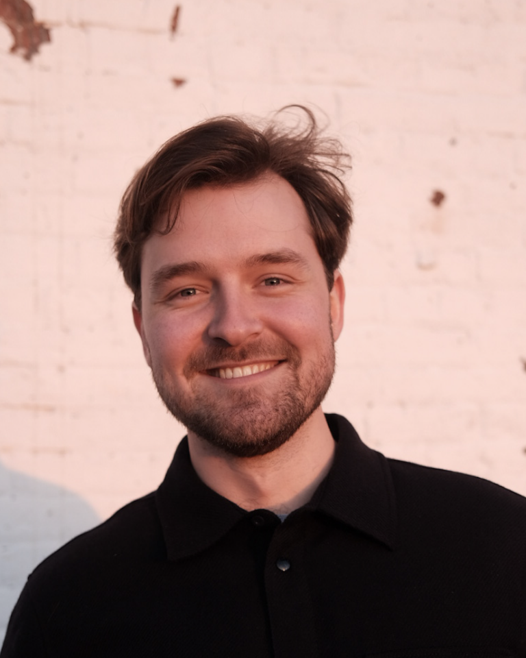 A man with brown hair and a beard smiles while standing in front of a white brick wall with some chipped paint, illuminated by warm sunlight.