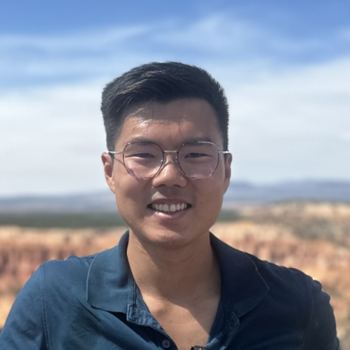 A person with short dark hair and glasses smiles outdoors, wearing a blue collared shirt. The background shows a blurred, sunny landscape with blue sky and reddish rock formations.