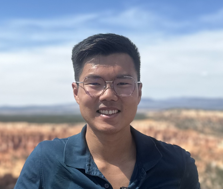 A person with short dark hair and glasses smiles outdoors, wearing a blue collared shirt. The background shows a blurred, sunny landscape with blue sky and reddish rock formations.