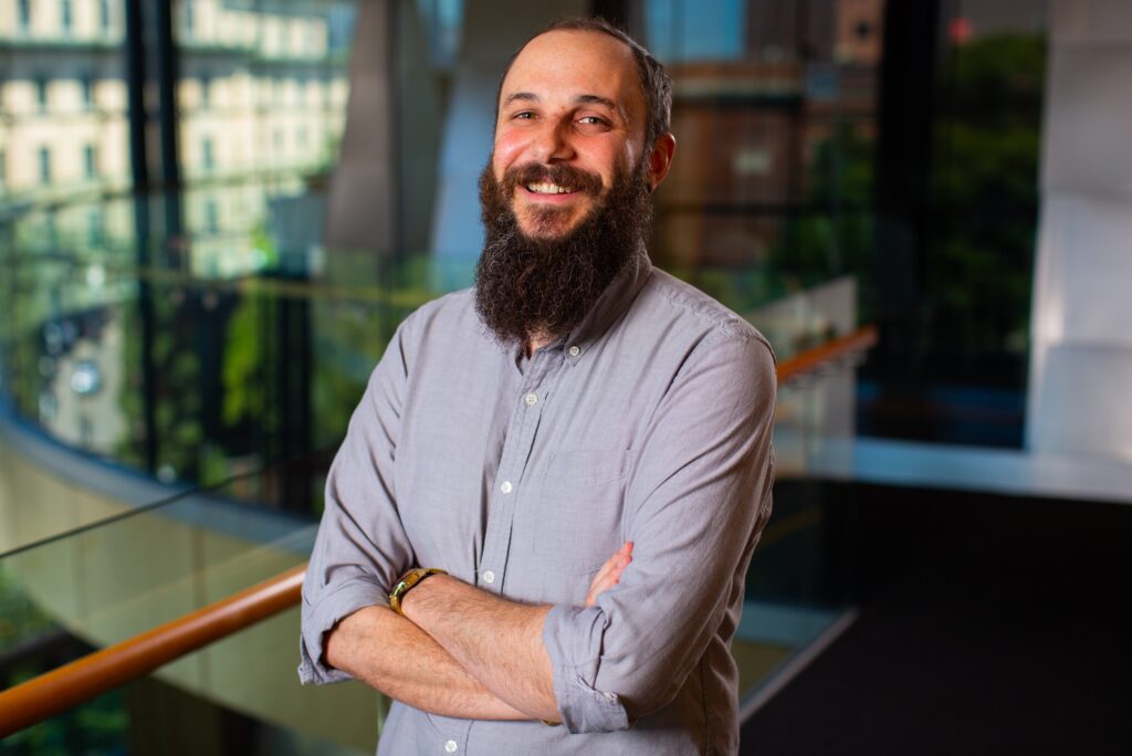 A bearded man with a gray shirt stands smiling with arms crossed in a bright, modern indoor space with large windows and a cityscape view in the background.