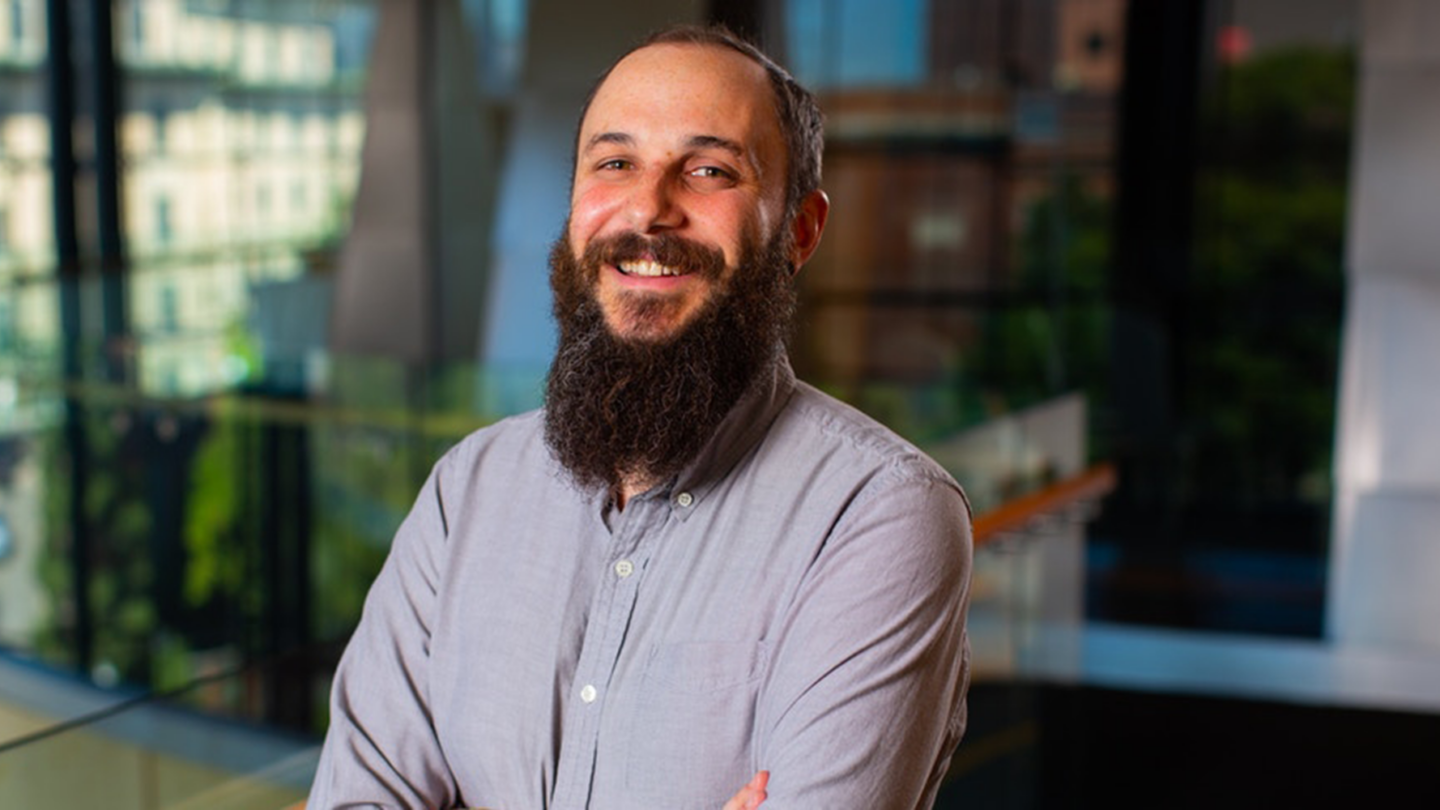 A man with a beard and mustache smiles with arms crossed, wearing a light gray button-up shirt. He stands indoors with large windows and blurred city buildings in the background.