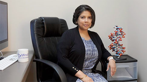 A woman sits in an office chair beside a desk, with a computer, a coffee mug, and a model of a DNA double helix on a drawer, looking at the camera.