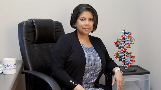 A woman sits in an office chair next to a desk with a DNA model and a mug. She wears a patterned dress with a black cardigan and looks at the camera.
