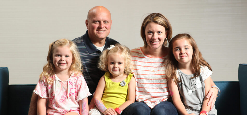A family of five sits on a blue couch, smiling. Two adults are behind three young girls, who are sitting in front, all facing the camera against a plain light background.
