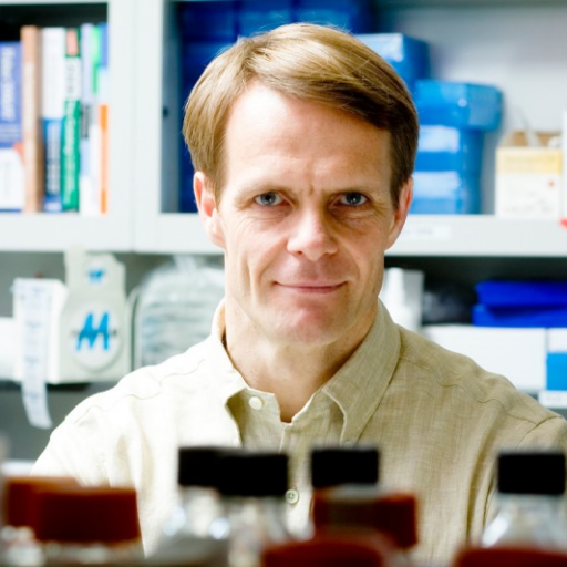 A man with light brown hair and a beige shirt smiles in a bright laboratory, surrounded by blurred lab equipment and shelves with books and supplies in the background.