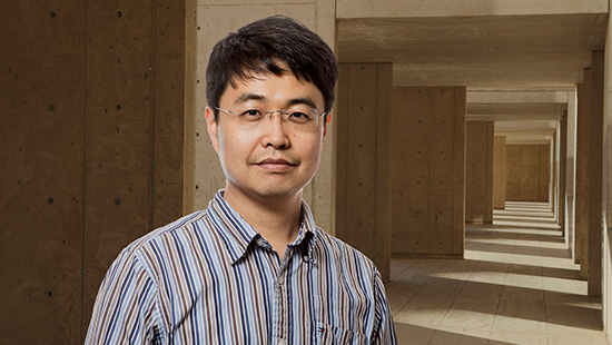 A man with glasses and short hair, wearing a striped shirt, stands in front of a background featuring concrete walls and columns with light streaming through.
