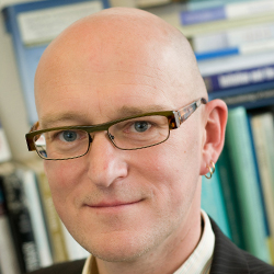 A person with a bald head, wearing rectangular glasses and a small hoop earring, is smiling softly. Shelves of books are blurred in the background.
