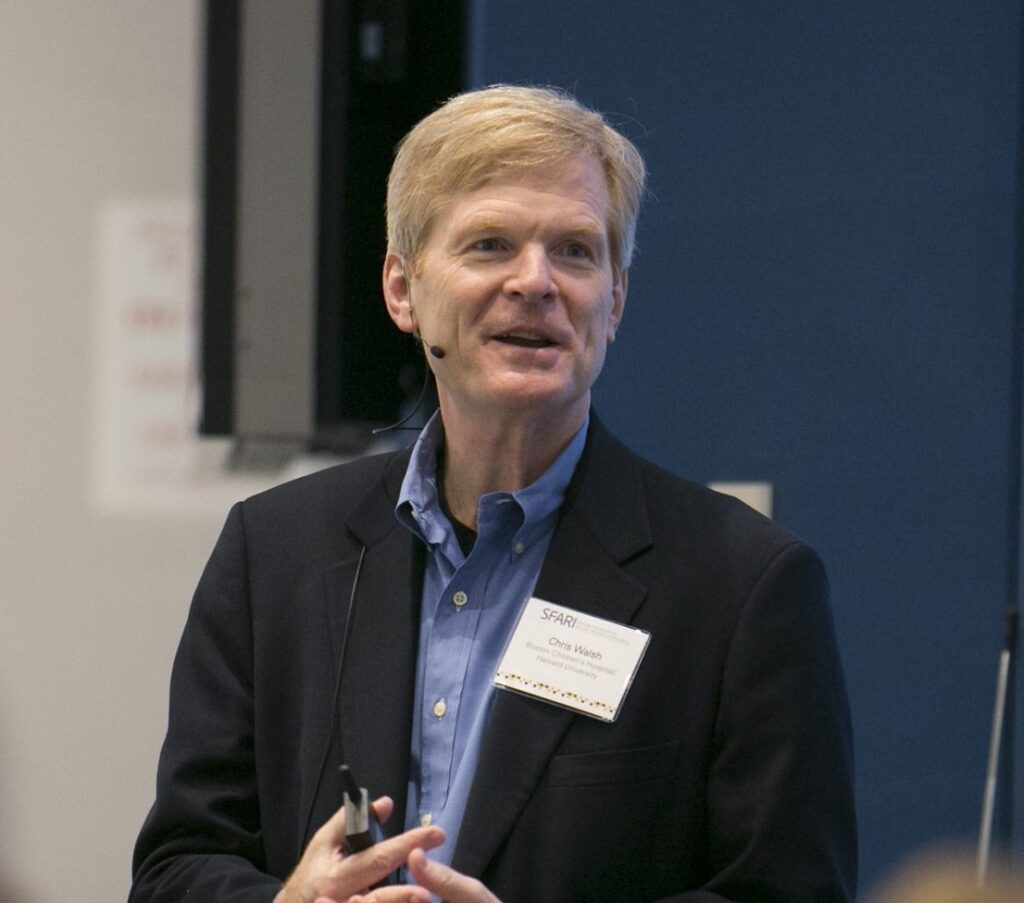 A man with blond hair in a dark blazer and blue shirt speaks, wearing a microphone headset and a name tag, standing indoors against a blue wall.