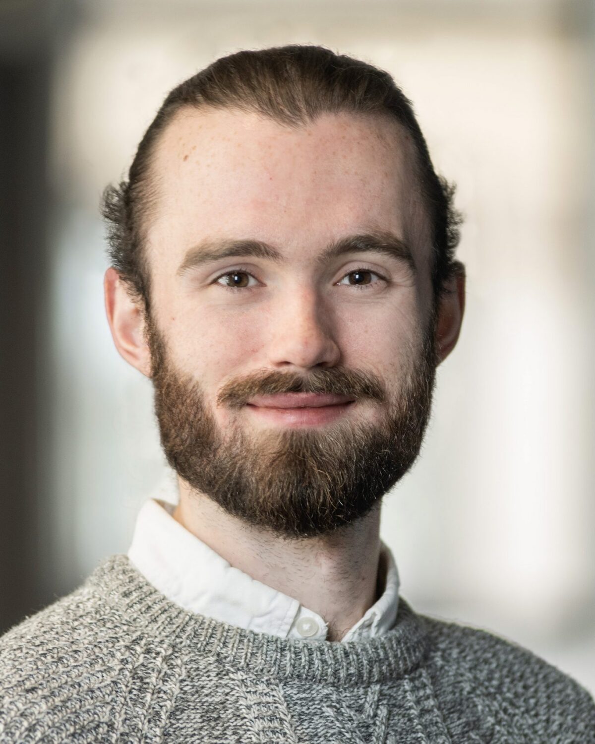 A man with a beard and mustache, wearing a gray textured sweater over a white collared shirt, smiles softly at the camera against a blurred indoor background.