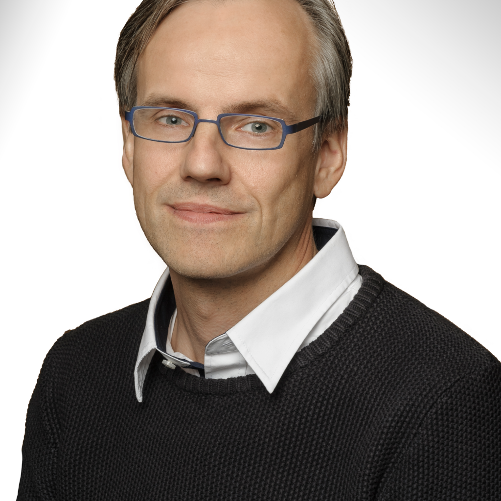 A person with light skin, short gray hair, blue glasses, and a neutral expression wearing a white collared shirt under a dark sweater, posed against a white background.
