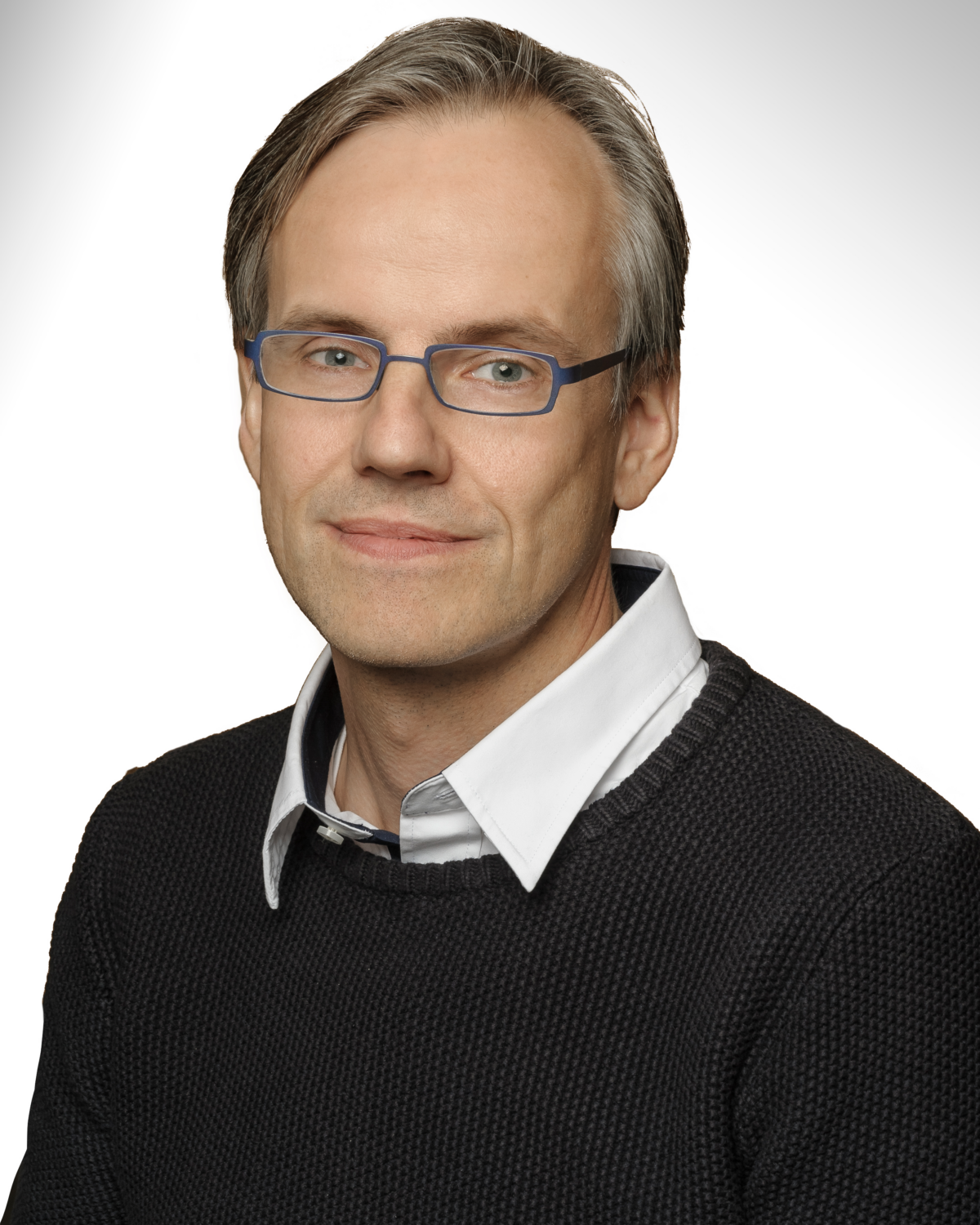 A person with light skin, short gray hair, blue glasses, and a neutral expression wearing a white collared shirt under a dark sweater, posed against a white background.