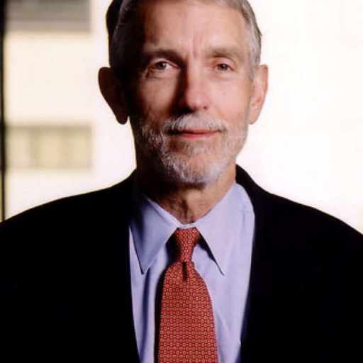 An older man with gray hair and a beard, wearing a dark jacket, blue shirt, and red patterned tie, stands in front of a blurred indoor background.