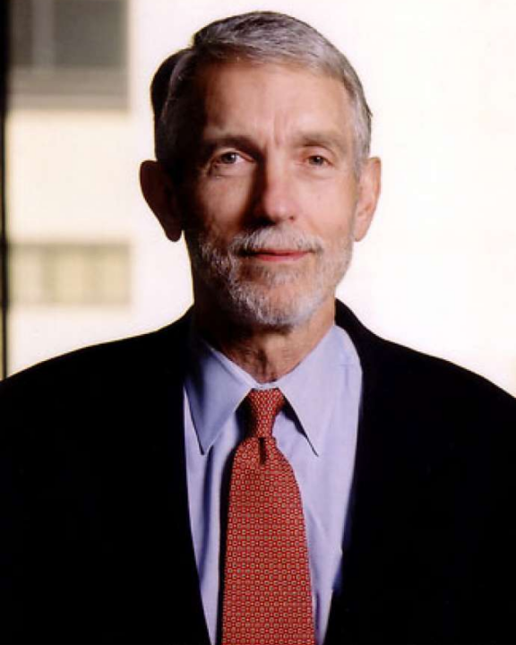 An older man with gray hair and a beard, wearing a dark jacket, blue shirt, and red patterned tie, stands in front of a blurred indoor background.