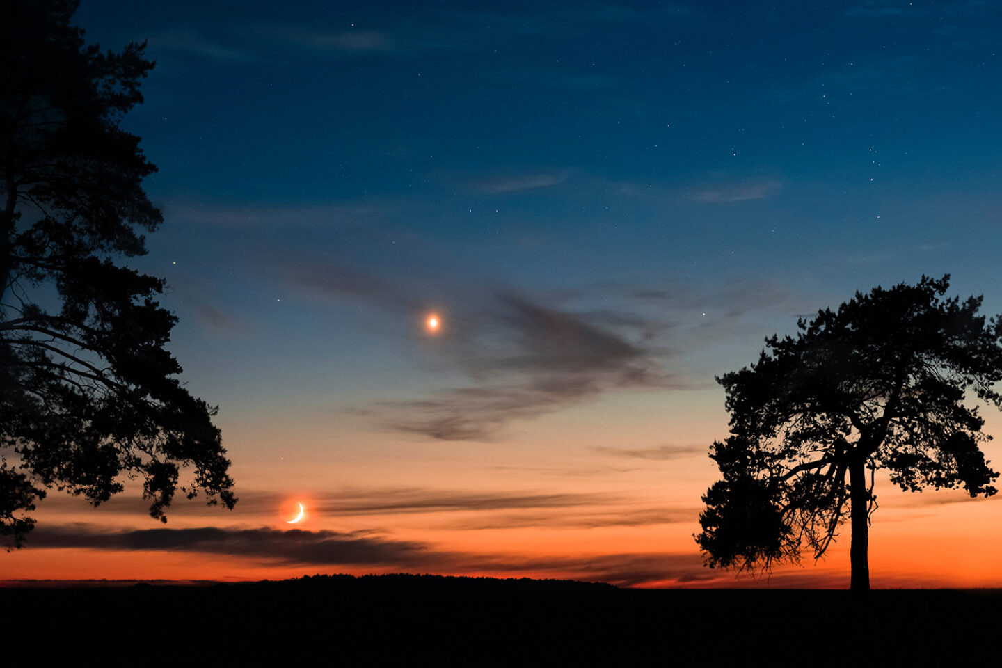 A serene sunset sky with a crescent moon and a bright planet visible, silhouetted trees in the foreground, and a vibrant gradient from orange to deep blue along the horizon.