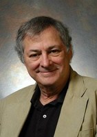 A man with gray hair wearing a tan blazer and black shirt, smiling at the camera against a neutral, softly lit background.