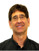 Man with short brown hair and glasses, wearing a dark collared shirt and smiling slightly, against a plain white background.