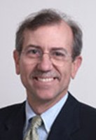 Portrait of a middle-aged man with glasses, short brown hair, and a suit with a light-colored shirt and tie, smiling in front of a plain light background.