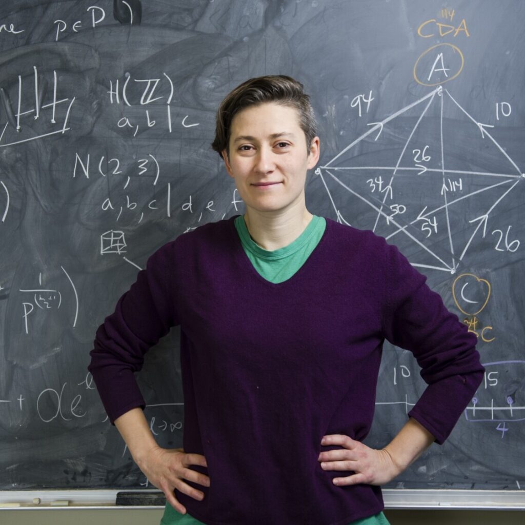 A person stands confidently in front of a chalkboard covered with mathematical equations, diagrams, and graphs. They wear a purple sweater and green shirt, with short hair, and hands on hips, looking at the camera.