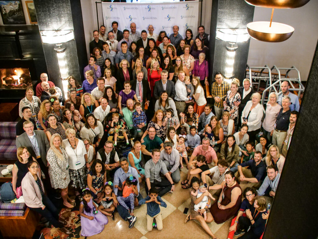 A large group of people of various ages and backgrounds pose together indoors for a group photo, smiling and looking up at the camera. Some adults hold children, and a fireplace is visible on the left.