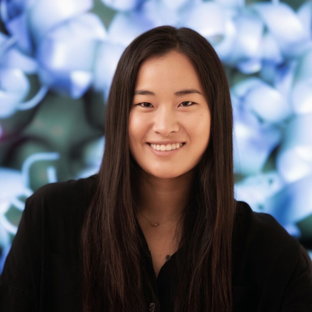 A woman with long dark hair smiles at the camera. She is wearing a black top and is posed in front of a blurred, abstract blue and white background.