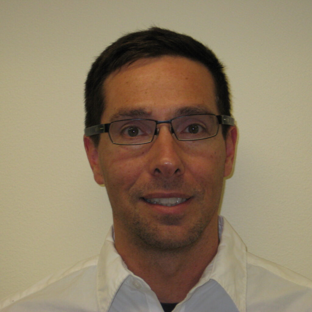 A man with short dark hair, wearing glasses and a white collared shirt, is smiling slightly against a plain light-colored background.