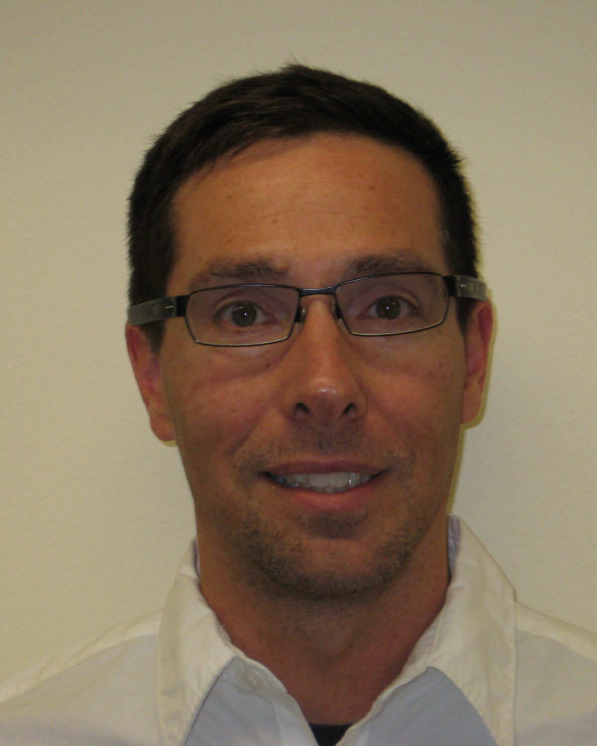 A man with short dark hair, wearing glasses and a white collared shirt, is smiling slightly against a plain light-colored background.
