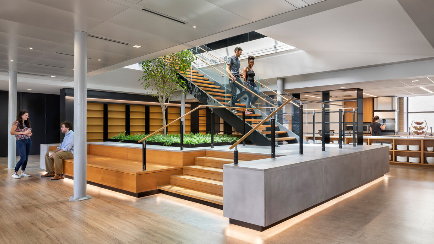 Modern office interior with a central staircase, indoor plants, and shelves. Four people are present: two walking upstairs, two chatting on the left. The space features wood accents, glass railings, and natural light.