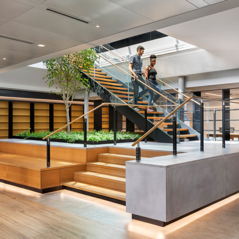 Modern office interior with a central staircase, indoor plants, and shelves. Four people are present: two walking upstairs, two chatting on the left. The space features wood accents, glass railings, and natural light.