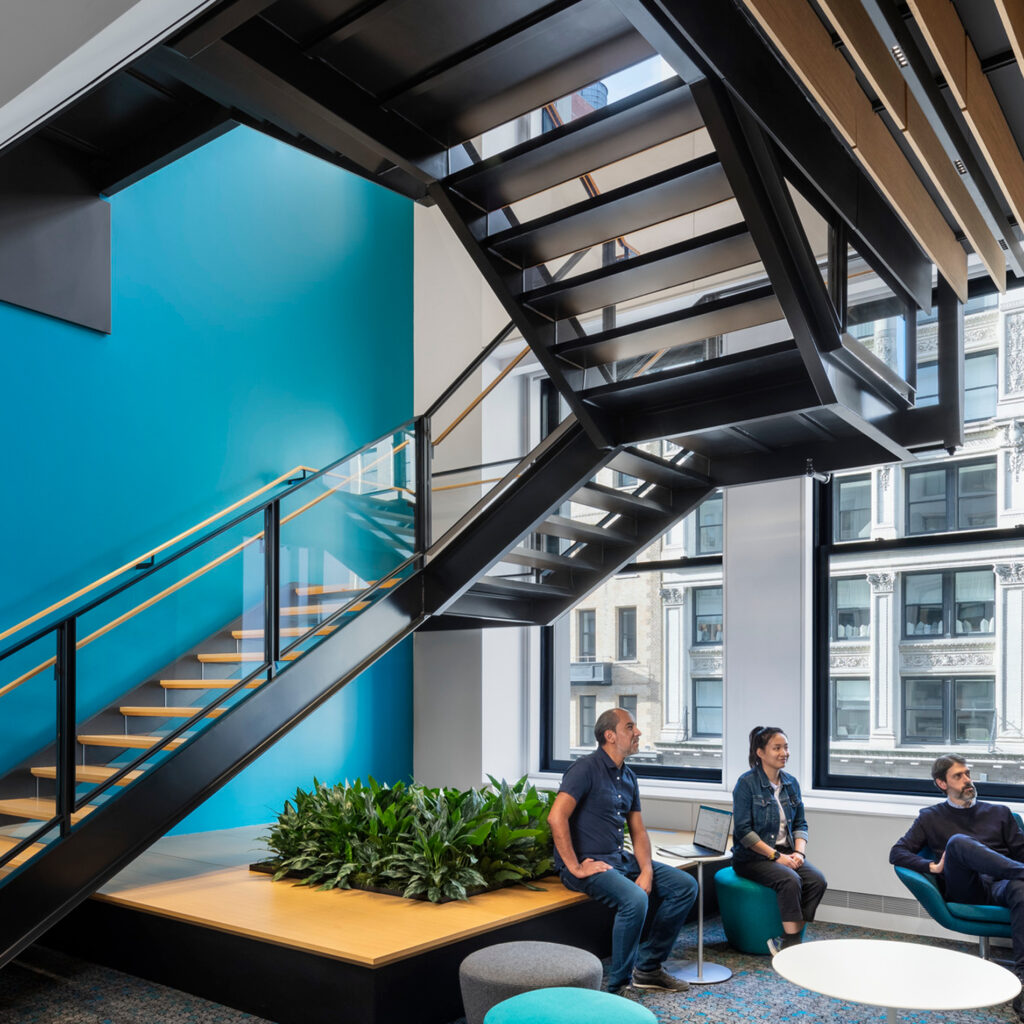 Three people sit near a modern black staircase in a bright office space with large windows, blue walls, indoor plants, and contemporary furniture.