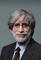 An older man with gray hair, glasses, and a beard, wearing a dark suit, light shirt, and patterned tie, smiles slightly against a plain, neutral background.