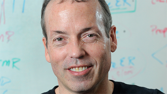 A middle-aged man smiling at the camera, wearing a black shirt, with a whiteboard covered in colorful marker writing in the background.