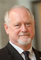 A middle-aged man with short white hair and a trimmed white beard, wearing a dark suit, light shirt, and patterned tie, is looking at the camera with a neutral expression.