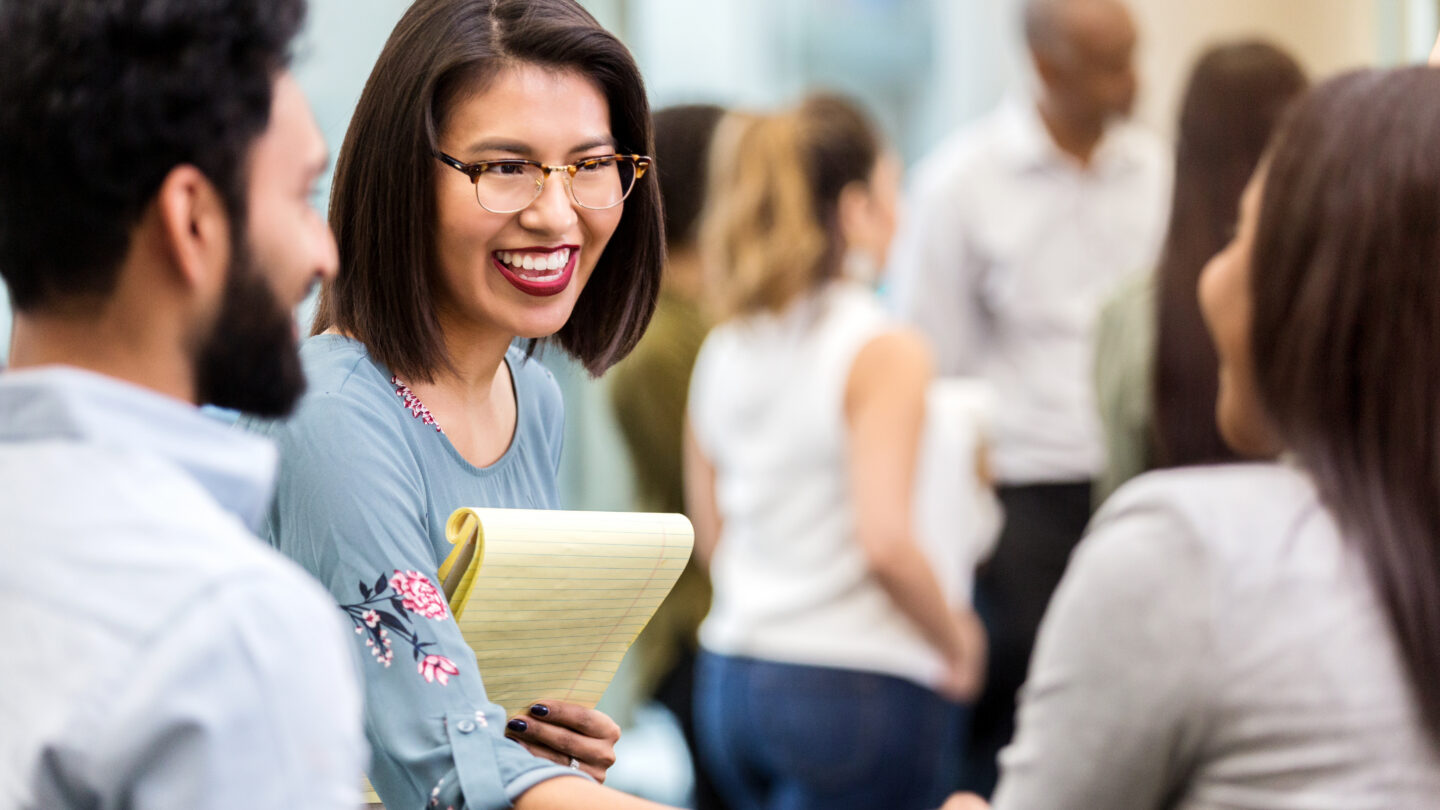 A cheerful young businesswoman stands with an unrecognizable coworker in a crowded office building and smiles as she shakes hands with her new unrecognizable client.