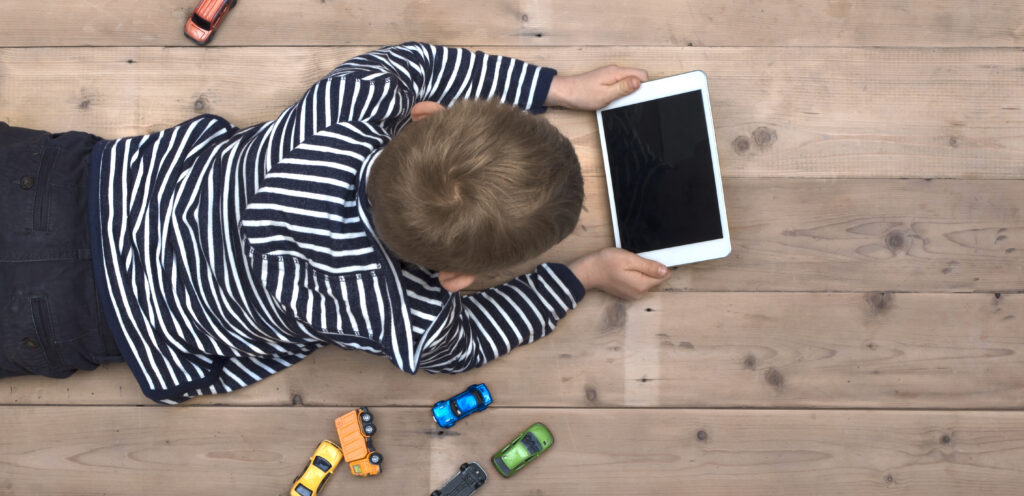 Photo of Top view header image with child laying down on the floor watching a movie on his tablet instead of playing with his toy cars